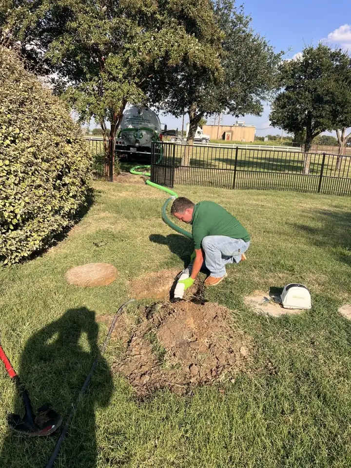 Diamond E crew member kneeling at a dug-out septic cleanout in a fenced yard, vacuum hose running to the truck